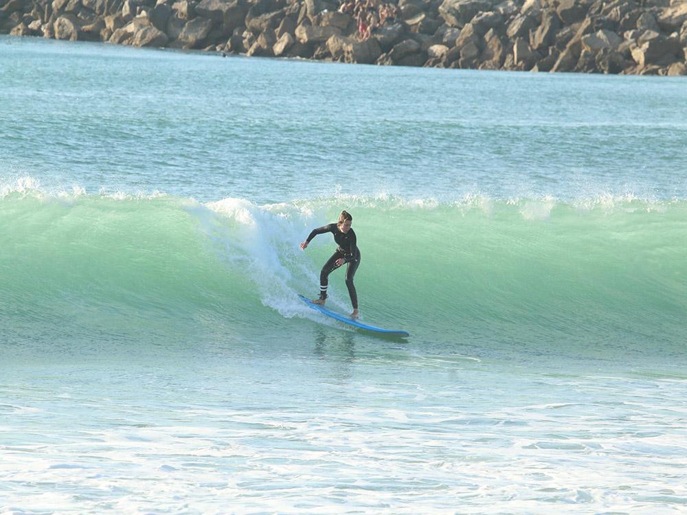 A surfer in a wetsuit riding down the face of a green wave with rocks in the background