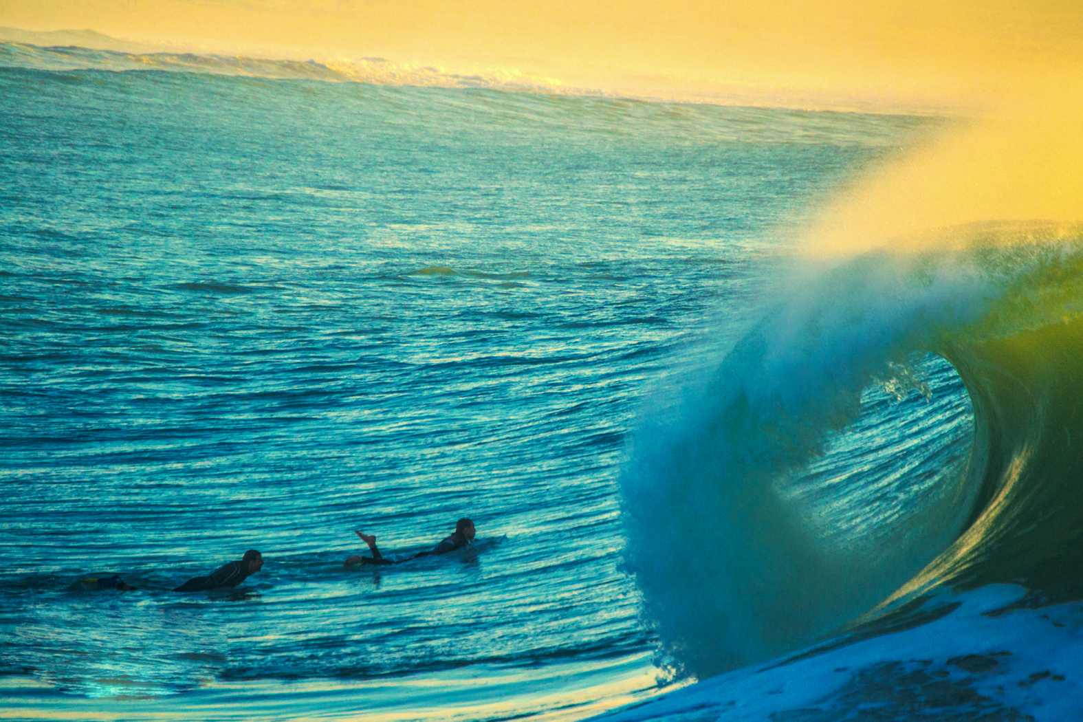Two surfers paddling toward a large curling turquoise wave at golden hour, dramatic warm and blue tones